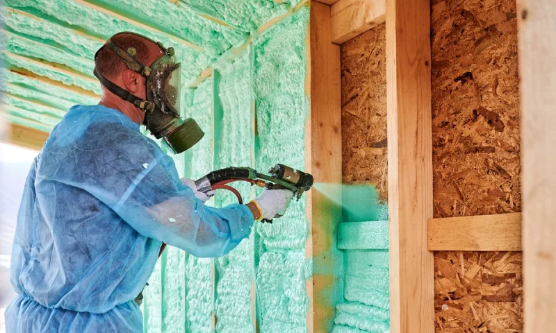 Certified applicator in a full-face respirator spraying foam into a wall cavity